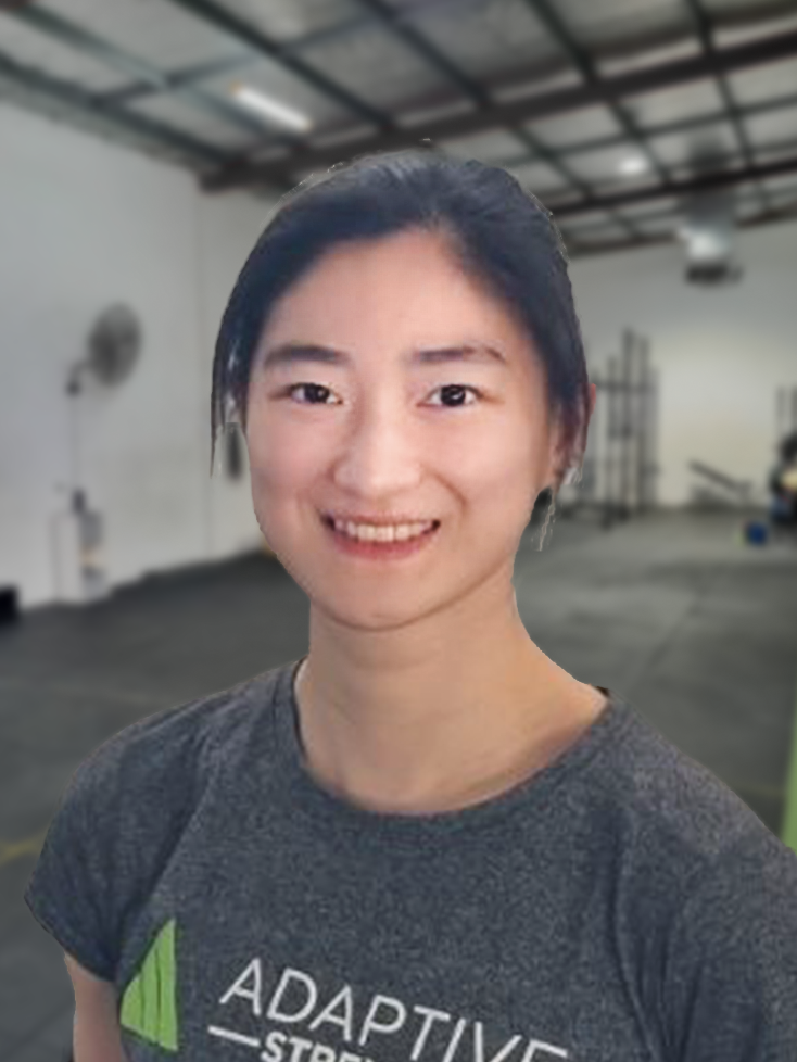 Smiling woman with dark hair wearing a gray shirt that reads ADAPTIVE, in a spacious indoor workshop setting.